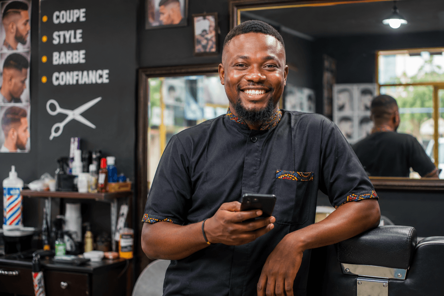 Coiffeur souriant avec son téléphone à la main, devant un mur affichant Coupe, Style, Barbe, Confiance et une icône de ciseaux